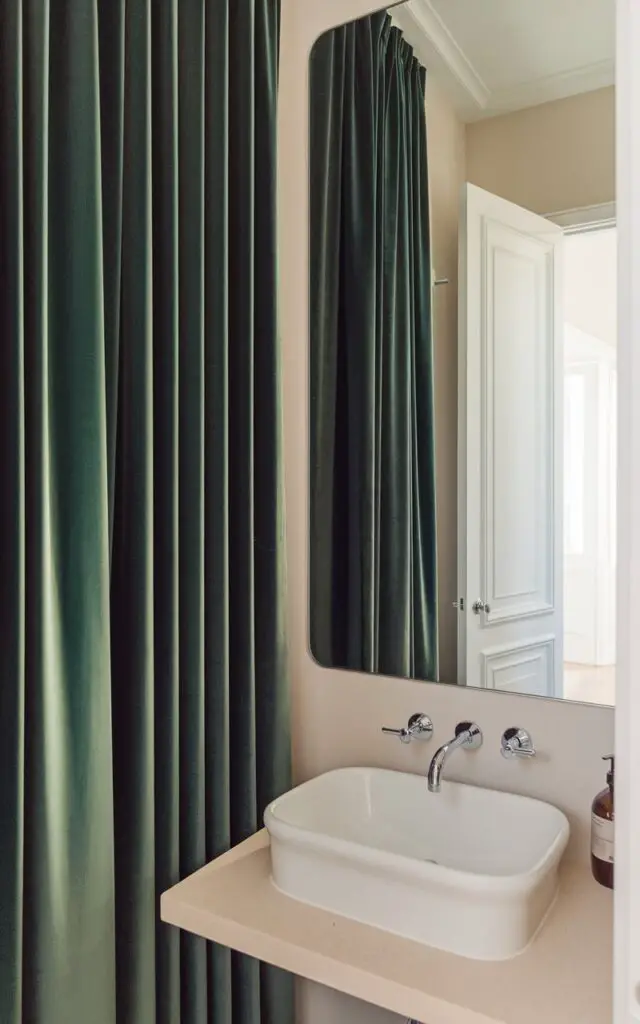 A photo of a small powder room with floor-to-ceiling drapes in deep emerald green framing the window. The drapes provide a touch of luxury and privacy. The fabric adds texture and warmth against the soft white walls and neutral floors. Below the window, a minimalist white sink is paired with a modern faucet in chrome. A simple, clear soap bottle sits on the countertop. A rectangular mirror reflects the bright open white door on the background, which shows an entryway, letting in natural light and amplifying the room's calm, elegant ambiance.