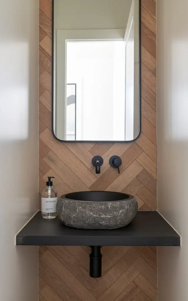 A photo of a small powder room with a herringbone wood accent wall. The wall runs behind a floating vanity with a stone basin and a minimalist modern faucet in matte black. A clear soap bottle with minimalist branding is placed on the corner of the vanity. The mirror, outlined in black metal, reflects the bright open white door in the background. The remaining walls are painted soft white.