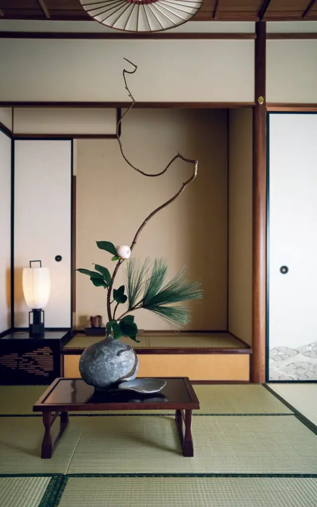 A refined Japanese living room with a striking ikebana arrangement on a vase. The arrangement features a curved willow branch, a single camellia bloom, and a sprig of pine. The vase is placed on a slender chabudai table. In the background, there is a Japanese low platform sofa upholstered in cream linen. The room has pale walls, a washi paper lamp, and wooden accents. The simplicity of the room allows the ikebana to shine, highlighting nature's grace and the beauty of restraint in design.
