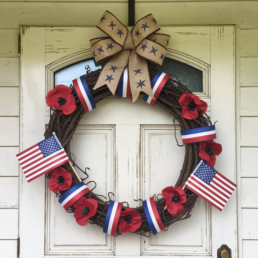 A photo of a white farmhouse with a wooden door. The door has a wreath made of intertwined grapevines wrapped in wide red, white, and blue ribbons. The wreath has miniature American flags and silk red poppies evenly spaced around the circle. At the top of the wreath, there is a big burlap bow with star accents.