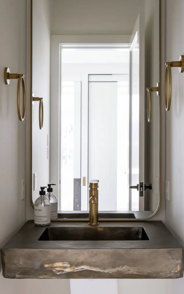 A photo of a luxurious small powder room with a metallic finish. The room features a brushed gold modern faucet sitting atop a stone countertop. There's a minimalist clear soap bottle with black text. Brass wall hooks and a gold-rimmed mirror add depth and glamour, contrasting with the matte white walls. The mirror reflects the bright open white door in the background, which reveals an entryway, allowing light to enter the room. The cohesive use of metallics elevates the powder room from functional to fabulous, blending elegance with a modern, minimalist approach.