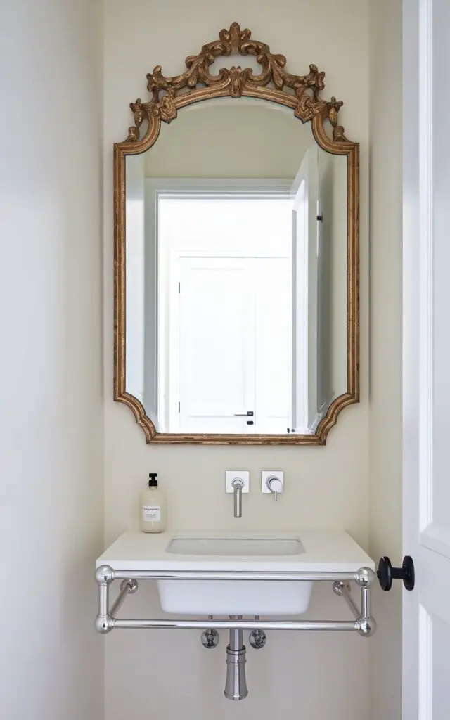A small powder room with a showstopping, oversized mirror with an ornate, gilded frame. The mirror is mounted above a slim floating vanity and reflects the bright open white door in the background. Below the mirror, a minimalist sink is paired with a sharp-lined modern faucet in brushed nickel. A clear soap bottle with a minimalist design rests neatly on the counter. The surrounding walls are soft ivory, allowing the dramatic mirror and metallic finishes to shine as the focal points in this clean, elegant room.