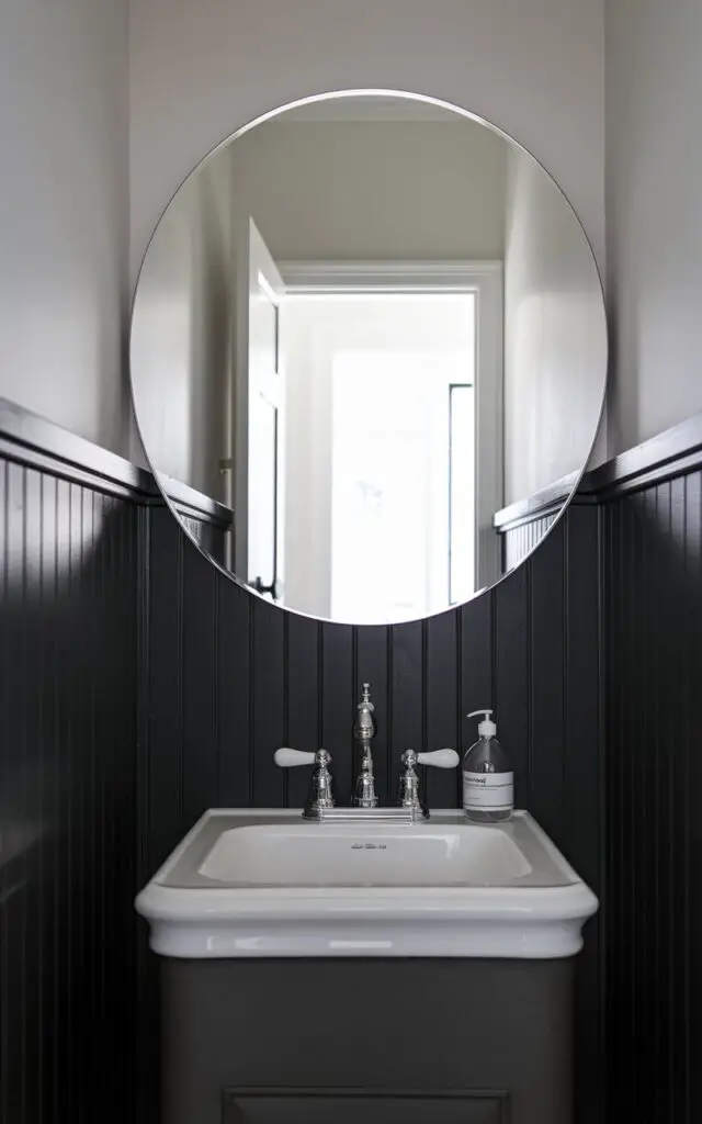A photo of a small powder room with rich wood paneling on the lower half of the walls. The paneling is painted a deep charcoal color, which contrasts beautifully with the soft white upper walls. This creates a sophisticated balance. A modern faucet in polished chrome flows from a sleek white sink placed atop a minimalist vanity. A clear soap bottle with a simple design sits next to the sink. Above, a large, round mirror reflects the bright open white door on the background, which shows an entryway, making the space feel more expansive. The paneling gives the room depth and character without overwhelming the design.
