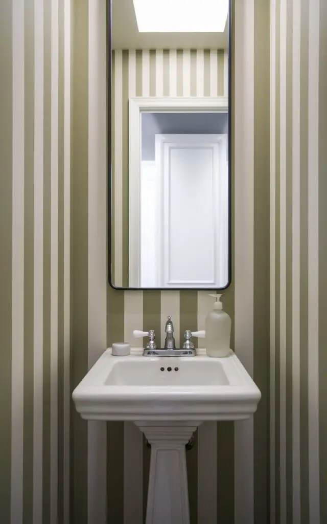 A photo of a small powder room with vertical beige and white stripes on the walls. The room contains a pedestal sink with a modern chrome faucet and a frosted glass soap bottle. There is a mirror with a thin matte black frame. The room has a bright open white door in the background. The overall effect is tall, tailored, and tastefully bold, with stripes lending structure to the compact space.
