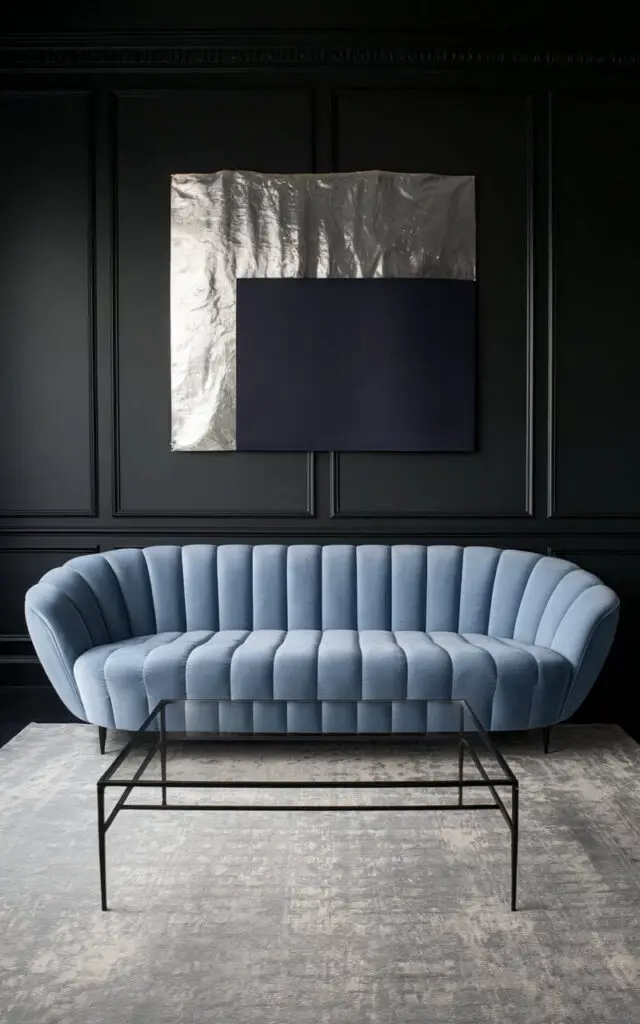 A dramatic blue and grey living room with deep charcoal grey walls. A cozy powder blue sofa is the centerpiece, providing a striking contrast against the dark backdrop. An oversized silver and midnight blue art piece hangs above the sofa, drawing the eye upward. A plain glass-top coffee table with black metal legs sits atop a pale grey rug, balancing the visual weight of the walls.