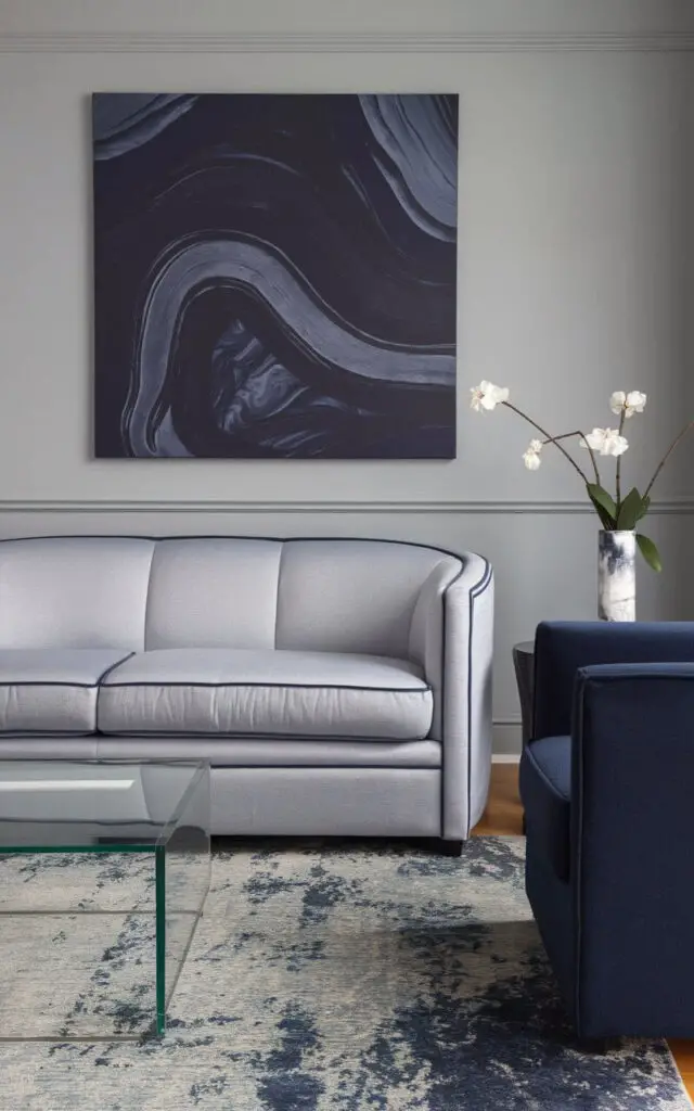 A photo of a refined blue and grey living room. There's a cozy sofa in soft dove grey with navy blue piping. There's also a navy blue armchair. The walls are a light fog grey. Above the sofa, there's a large canvas with abstract swirls in navy and ash. In front of the sofa, there's a plain glass coffee table. Underneath all of this, there's a rug with dusty blue and charcoal tones.