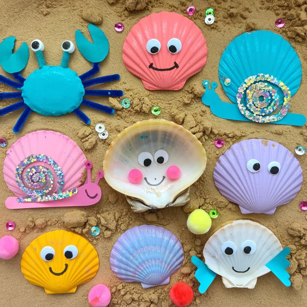 A homemade craft for kids featuring various seashell critters arranged on a sandy surface. Each shell has been painted in bright, cheerful colors like turquoise, coral pink, sunny yellow, and lavender. The creatures include a painted blue crab with pipe cleaner legs, a glittery snail with googly eyes, and a clam shell transformed into a smiling fish with paper fins. The shells vary in size and texture, creating visual interest. Some shells are decorated with sequins, beads, and mini pom-poms.