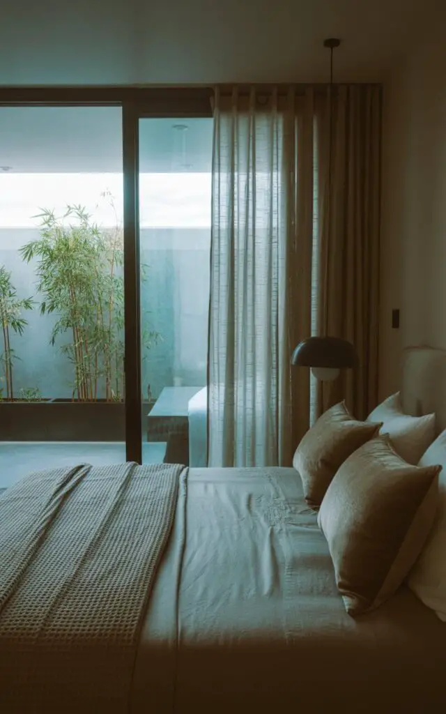 A photograph of a serene modern bedroom bathed in soft, natural light. The room centers on a low-profile bed draped in cool-toned linens, a textured waffle blanket, and plush velvety pillows, positioned directly beneath a large window with sleek sliding glass panels. The window is partially open, revealing a quiet patio with potted bamboo and sheer curtains gently rippling in a soft breeze; a matte black pendant lamp hangs beside the bed, subtly illuminating the scene. The overall ambiance is one of minimalist elegance and effortless relaxation, with a calming color palette and clean lines contributing to a sense of tranquility.