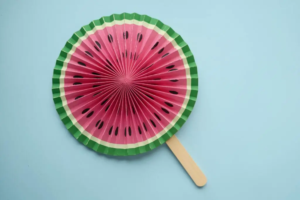 A photo of a handcrafted fan-like object that resembles a slice of watermelon. The fan is predominantly pink with black seeds and has a green border. The fan is mounted on a wooden stick. The background is a light blue shade.