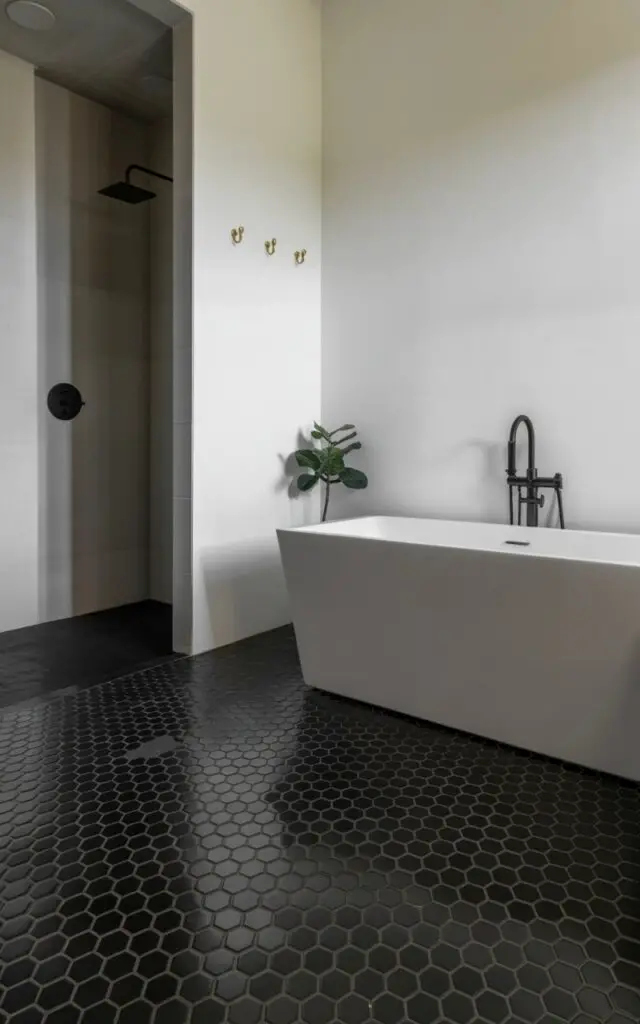 A photograph of a sleek, modern bathroom showcasing a striking black hexagon tile floor. The matte black tiles create a bold geometric pattern that extends across the floor, contrasting beautifully against bright white walls, with a freestanding white bathtub taking center stage in the composition. To the left, a minimalist shower cube with black fixtures reflects soft ambient light, while brass towel hooks add warmth, and a single leafy green plant sits in a corner. The entire scene is bathed in soft, diffused lighting, accentuating the bathroom's sophisticated and edgy design.