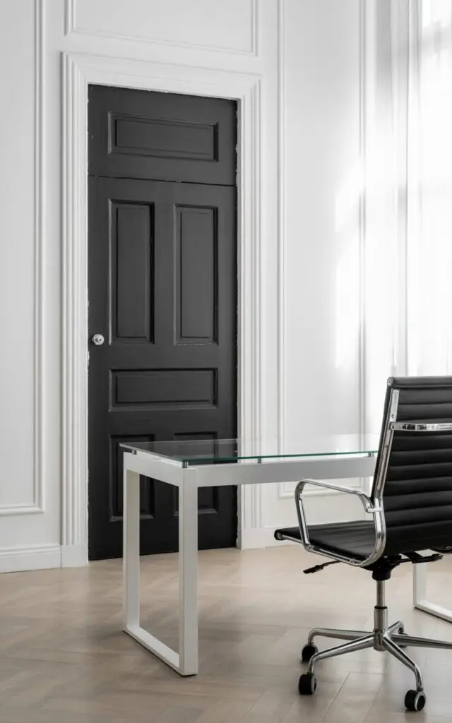 A photograph of a modern black and white office centered around a striking matte black paneled door. The door stands prominently against clean white walls and light-toned wooden flooring, its deep black finish providing a bold contrast. In front of the door, a minimalist white desk with a glass top is paired with a black ergonomic chair featuring chrome accents and comfortable padding, both perfectly aligned. Soft, diffused light streams in from a large window to the side, highlighting the monochrome gallery wall and creating a sense of quiet sophistication.