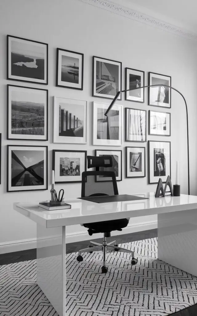 A black and white photo of a stylish office with a gallery wall of black and white photography. The office has a clean and minimalistic design, with a bright white lacquer finish table and a black mesh chair. The photographs range from sweeping landscapes to close-up architectural details, each framed in thin matte black for uniformity. The clean white wall allows the gallery to shine, while a soft black-and-white woven rug adds comfort underfoot. A slender floor lamp with an arched black stem complements the visual flow. Every detail contributes to a very well decorated workspace rich in artistry and sophistication.