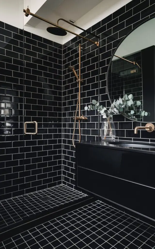 A moody bathroom with charcoal-colored bathroom tiles. The tiles cover the shower wall cubicle and floor, creating a seamless and immersive dark palette. The bathroom has brushed gold fixtures and a large circular mirror. There's a floating white vanity and a clear vase with eucalyptus sprigs. The charcoal tile sets a bold, confident tone, making the space feel like a high-end hotel suite.