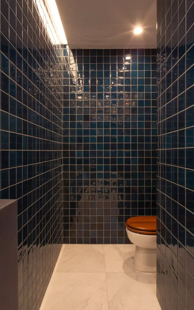 A photograph of a luxurious bathroom dominated by deep navy blue tiles. The walls are meticulously covered in the navy tiles, contrasted by crisp white grout lines and reflecting a soft glow from the recessed lighting above. A pristine white marble floor extends across the room, culminating in a glimpse of a modern white toilet with a wooden seat. Gentle, diffused light highlights the textures of the tile and marble, creating a serene and sophisticated atmosphere.