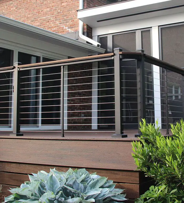 A photo of a sleek balcony with horizontal metal bars that stretch across a low-profile frame. The balcony railing is well designed with evenly spaced tubular bars, precisely aligned with laser-cut end caps, creating a confident linear rhythm. The posts are square-sectioned steel, powder-coated in matte charcoal for weather resistance. Bar spacing is measured to safety code and visually generous. The top rail has a slight ergonomic curve for comfortable leaning. The balcony sits against a white stucco wall. Morning light forms thin shadows of the bars across the decking, emphasizing the structure's linear geometry and modern appeal.