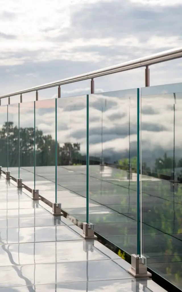 A photo of a balcony with a seamless tempered glass railing. The railing appears to float as there are no visible posts. The glass panels are anchored into the floor with hidden channels, keeping the edges polished and smooth. The railing has a slim stainless-steel cap at the top, which gleams subtly in the sun. The glass panels reflect the clouds and greenery, enhancing the ethereal quality of the scene. The overall effect is airy, modern, and luxurious, blending safety with an uninterrupted connection to the outdoors.