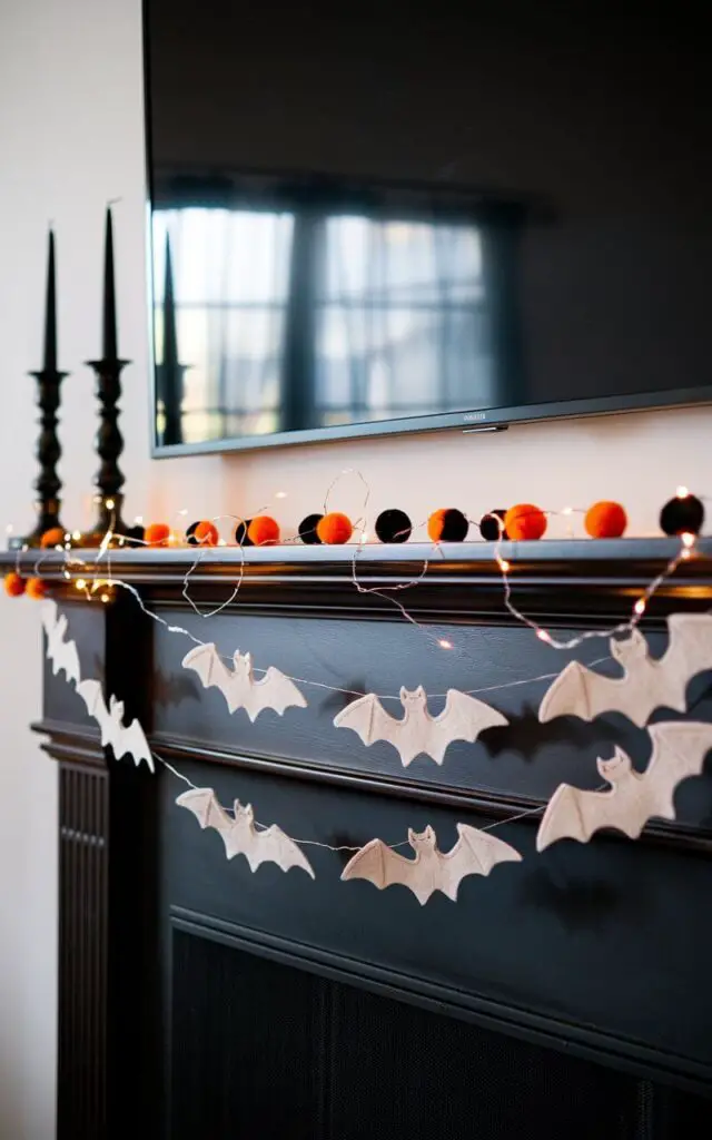 A well-designed mantel decorated with a layered Halloween garland. One garland is made of felt bats, their wings casting playful shadows. The other features orange-and-black pom-poms. Twinkle lights twist through the garlands, glowing softly against the dark wood of the fireplace. The Halloween mantel feels festive yet minimalist. A pair of black candlesticks at either end provides balance. An oversized black television is mounted above the mantel.