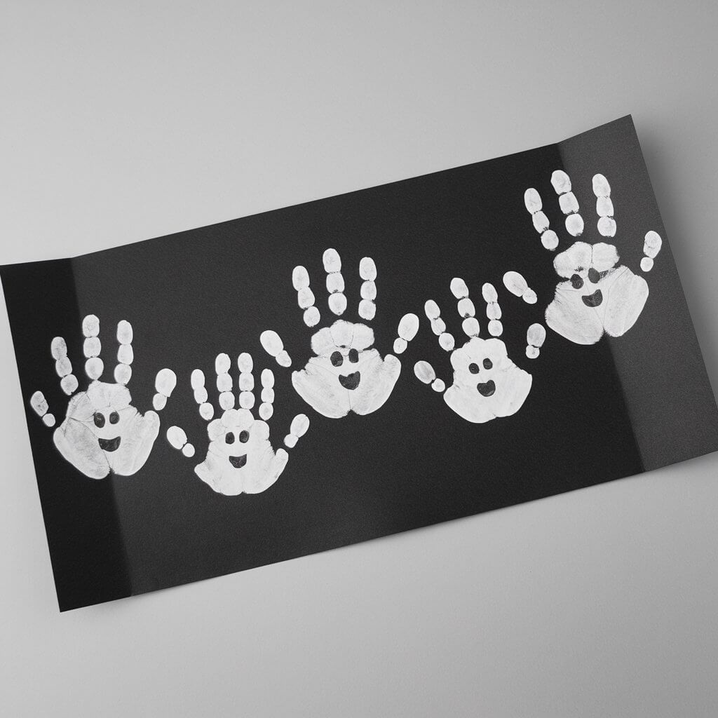 A minimalist Halloween-themed craft of white handprint ghosts painted onto solid black construction paper. The ghosts have the full palm shape with fingers stretched out, resembling floating spirits. Black dots form the eyes and small ovals mark the mouths, giving each ghost a slightly different spooky expression. The ghosts are neatly aligned on the page, creating a family of handprint characters. The artwork is lying flat, placed on a plain table, and there are no tools visible. Only the finished artwork is in the frame.