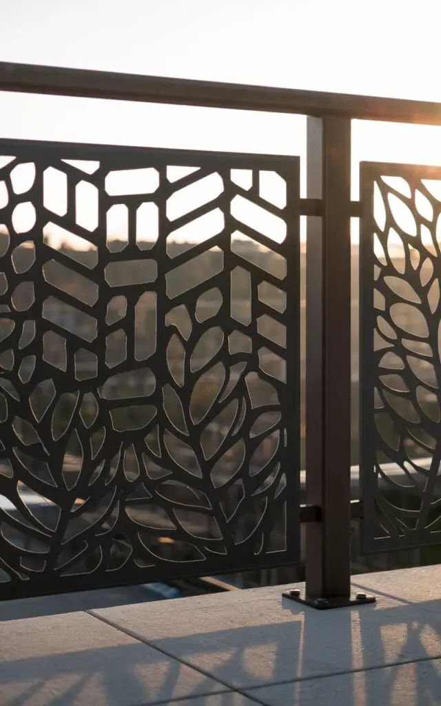 A photo of a contemporary laser-cut metal panel serving as a balcony railing. The panel is powder-coated steel with a geometric pattern of hexagons and stylized leaves. The railing has a flat top rail and a flush base. The panel is mounted with hidden clamps behind a vertical post. In the late afternoon, sunlight streams through the cutouts, casting patterned shadows onto the balcony floor.