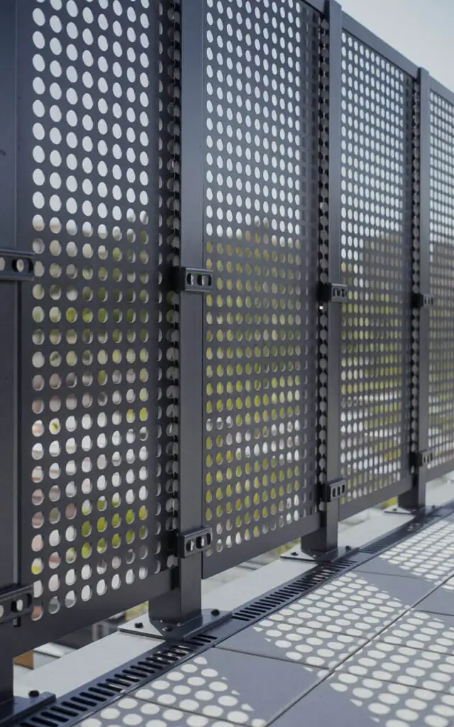 A photo of a balcony railing made of perforated steel sheets with uniform circular cutouts, forming a grid. The railing is laser-cut, powder-coated in matte charcoal, and bolted to structural posts with seamless brackets. Drainage slots are present at the base for weather protection, and the edges are chamfered for safety. Light filters through the holes, casting intricate dot patterns onto the balcony tiles below. The railing provides privacy without blocking air circulation, all in a clean, contemporary style.