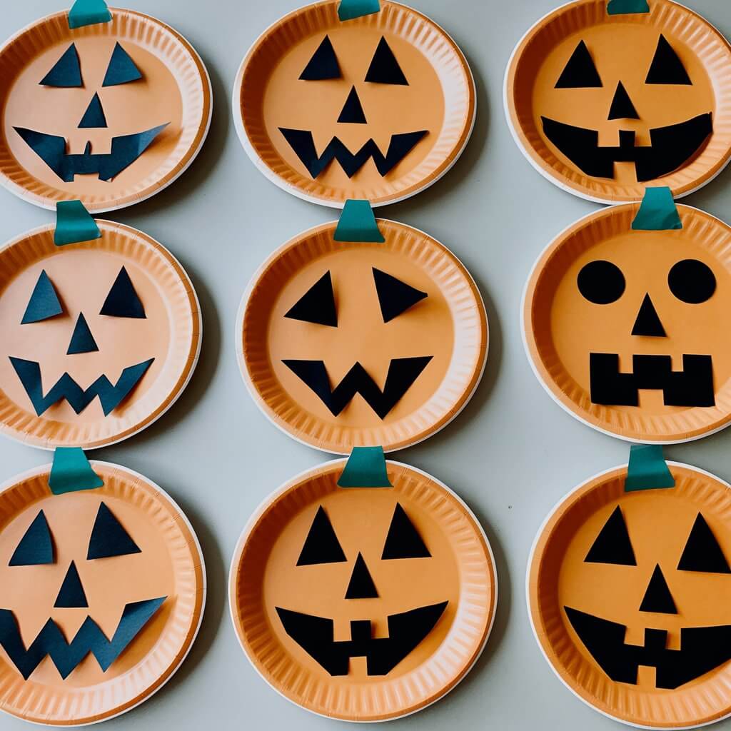 A minimalist photo of a table with several orange paper plates decorated to look like jack-o'-lanterns. Each plate has a different Halloween face, with some having triangle eyes and jagged mouths, and others having big goofy grins. The plates are placed neatly on the table, with no additional tools or decorations visible.
