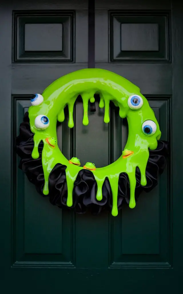 A photo of a Halloween wreath hanging on a dark forest-green door. The wreath is made of fake slime with neon green gel-like drips trailing over the edges. There are plastic eyeballs and small frogs peeking from beneath the slime. The base of the wreath is wrapped in matte black fabric. The entryway is minimalist, with no additional props. The lighting is controlled, highlighting the gloss of the slime and emphasizing the contrast without visual clutter.