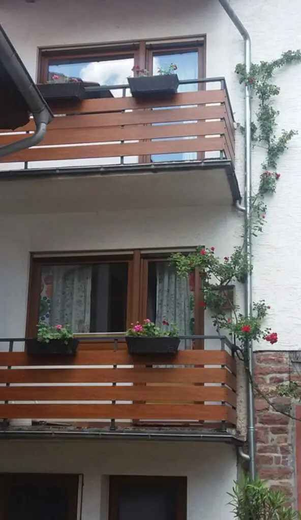 A close-up photo of a balcony railing made from natural wood slats. The railing is made of seasoned teak wood with an oil finish and has rounded edges for safety. The wood is held in place by stainless-steel spacers and is anchored to concealed steel posts. The railing has a water-repellent sealer that highlights the grain and protects against weathering. The railing has a slim metal top cap and a drip edge. The railing has a warm, tactile, and impeccably detailed appearance.