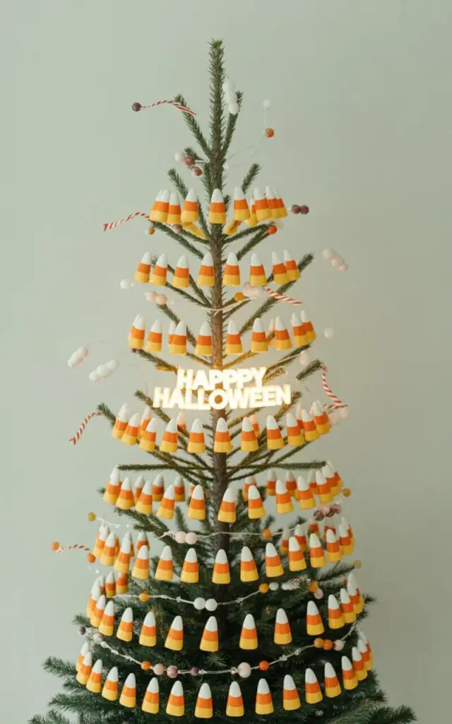 A photograph showcasing a slender fir tree meticulously decorated in a Halloween candy theme. The tree is adorned with ornaments arranged in precise, horizontal bands of white, orange, and yellow, mimicking candy corn, with delicate faux candy pieces and thin striped ribbon sparingly draped across the branches.  A single string of "Happy Halloween" lights is interwoven throughout the branches, casting a warm, diffused glow onto the tree and highlighting the layered candy design. The tree stands centered against a soft, pale gray backdrop, creating a clean and playful aesthetic.