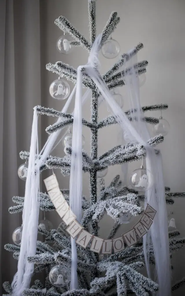A photograph of a minimalist Halloween tree standing in a softly lit room. The tree features a flocked white structure, meticulously adorned with sheer gauze strips that suggest drifting spirits, and sparsely hung transparent baubles displaying faint ghost faces. Subtle frosted ornaments and silver details are woven among the branches, catching the light and enhancing the ethereal aesthetic, while a single "Happy Halloween" banner is draped gently across a lower branch. The backdrop is a soft gray, creating an uncluttered, ghostly effect and highlighting the tree's serene and otherworldly presence.