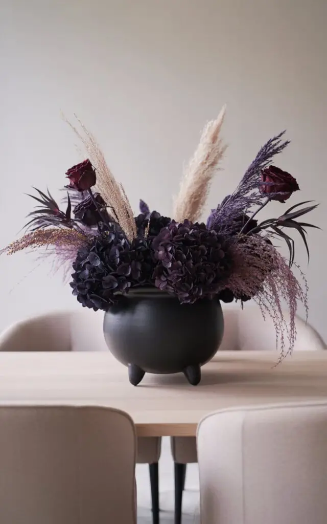 A photograph of a meticulously arranged centerpiece showcasing a matte black cauldron filled with a captivating mix of dark purple hydrangeas, soft pampas grass, and dried burgundy roses. The cauldron rests on a clean, minimalist dining table, where the florals spill naturally and elegantly over its rim, creating a visually appealing overflow. Light taupe upholstered dining chairs are subtly positioned in the background, providing a soft contrast to the dark centerpiece. Soft, diffused lighting highlights the textures and colors of the arrangement, creating a chic and balanced Halloween aesthetic.