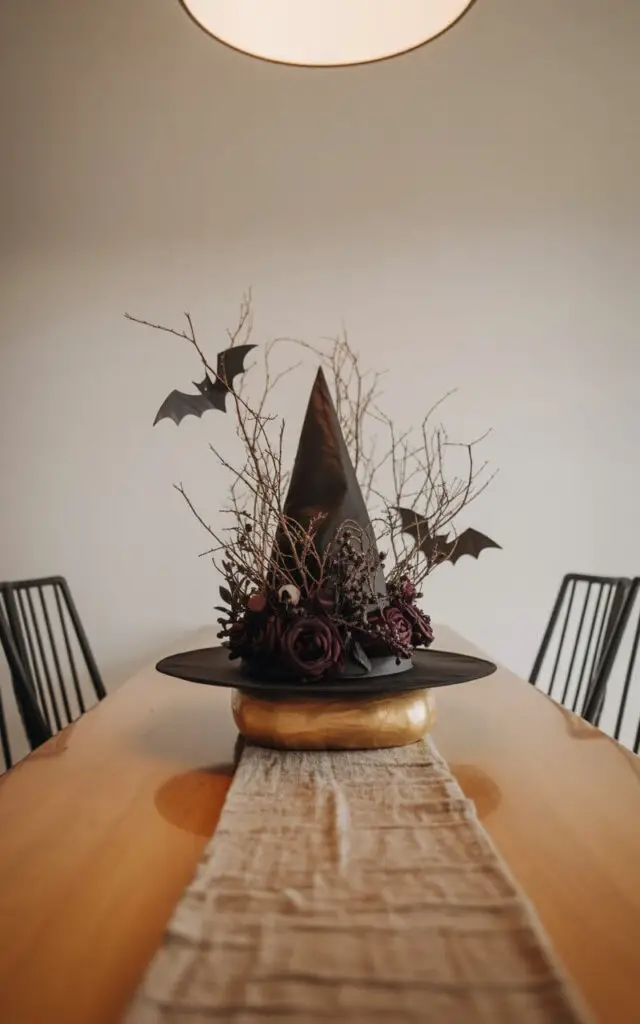 A photograph of a sophisticated Halloween centerpiece featuring an upside-down black witch hat filled with a dramatic arrangement of dried branches, deep burgundy dark red roses, and delicate faux bats. The hat sits atop a smooth, circular wooden base centered on a clean, minimalist dining table, spotlighted by a soft overhead light. Simple black metal dining chairs frame the centerpiece without distraction, while a textured linen runner subtly accents the table. The lighting is warm and diffused, creating a chic and understated seasonal atmosphere.
