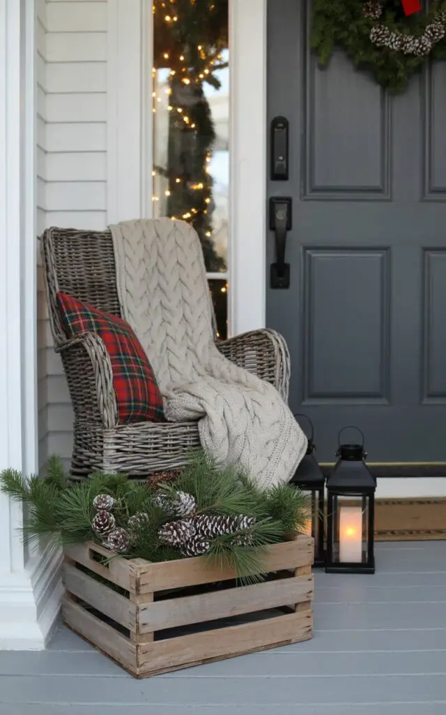 A photograph of an elegantly minimalist Christmas front porch showcasing carefully curated seasonal decorations against clean architectural lines. A weathered wicker chair holds a chunky cream cable-knit throw draped over a red and green plaid cushion, positioned beside a rustic wooden crate overflowing with fresh pine boughs and silver-dusted pinecones. The charcoal gray front door features classic black hardware and is flanked by matching black metal lanterns casting a warm amber glow. Delicate evergreen garland with subtle white berries frames the doorway, while the layered textures of natural wood, woven materials, and fresh greenery create an inviting yet sophisticated holiday welcome under soft winter daylight.
