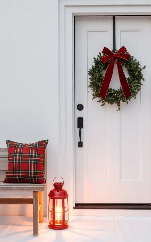 A photograph of a minimalist Christmas-decorated front porch featuring clean lines and vibrant holiday colors against a pristine white backdrop. A simple evergreen wreath adorned with a luxurious red velvet bow hangs centered on a white door with a sleek black handle and traditional keyhole. To one side, a wooden bench displays a single red and green plaid throw pillow, while a glowing red metal lantern casts warm golden light nearby. The scene captures the essence of modern holiday elegance with uncluttered composition, soft natural lighting, and the perfect balance of classic Christmas red and green against crisp white architectural elements.
