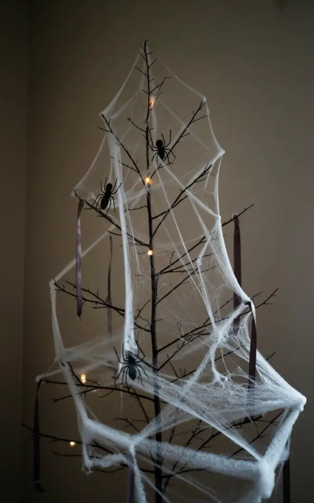 A photograph of a lone Halloween tree standing in a softly lit, darkened room. The tree, adorned in a minimalist spooky style, is meticulously wrapped in fine layers of faux spiderwebs strategically placed to avoid visual clutter, with a few large, realistic-looking spiders spaced evenly along its branches. Subtle LED lights embedded within the webbing gently highlight the delicate structure without overpowering the tree’s silhouette, while dark purple ribbons peek through in measured lengths, adding a touch of elegance. The background remains intentionally uncluttered and dim, allowing the tree to become the sole focus of the image.