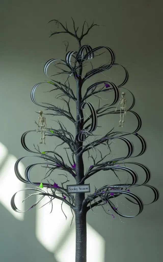 A stylized illustration of a Halloween tree, evoking a gothic whimsical aesthetic, stands centered against a muted gray wall. The tree's branches are meticulously adorned with black and white striped ribbon forming precise, elegant spirals, complemented by crooked twig accents and strategically placed skeleton figurines. Subtle pops of deep purple and a touch of neon green illuminate the spiraling ribbon and skeletal figures, adding depth and contrast to the dark branches. Soft, diffused light from an unseen source casts gentle shadows, highlighting the tree’s eerie elegance and Burton-inspired symmetry in a darkly charming presentation, with the tree’s label reading “Spooky Season”.