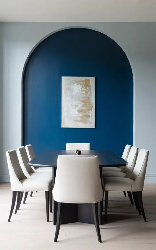 A photograph of an elegant modern dining room featuring a striking indigo accent wall that serves as the room's dramatic focal point. In front of the wall sits a sleek rectangular dining table with a smooth matte black finish, surrounded by six classic dining chairs upholstered in creamy off-white linen fabric. A single piece of contemporary abstract artwork in muted gold and white tones hangs centered on the indigo wall, while the remaining walls are painted in soft dove gray. Natural light from large windows illuminates the space, creating gentle shadows and highlighting the sophisticated contrast between the deep navy blue accent and the lighter neutral furnishings.