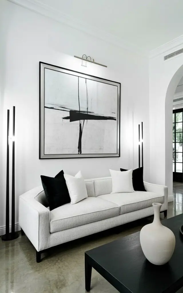 A low-profile modern sofa upholstered in a soft gray-white fabric sits centered in the room. Above the sofa, a large abstract artwork with a white background and black geometric shapes hangs on the white wall, framed by a thin black metal frame. A slender black coffee table positioned in front of the sofa holds a sculptural vase with a smooth, matte finish and a gently curved silhouette. Tall, narrow black floor lamps stand on either side of the sofa, casting subtle shadows across the polished concrete floor and the surrounding empty space.
