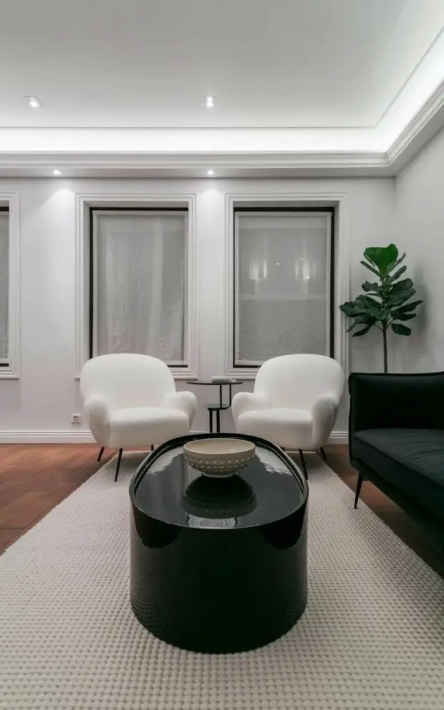 Two plush white boucle chairs with rounded backs and thin black legs face a sleek black sofa upholstered in a smooth fabric. A black oval coffee table with a polished surface sits centered between the chairs and sofa, displaying a large ceramic bowl with a matte finish and a geometric pattern. A thick, off-white rug with a subtle woven texture covers the hardwood floor, while recessed lighting fixtures are integrated into the ceiling. Large windows reveal a muted gray cityscape visible through sheer linen curtains, and a single potted fiddle-leaf fig plant stands in the corner.