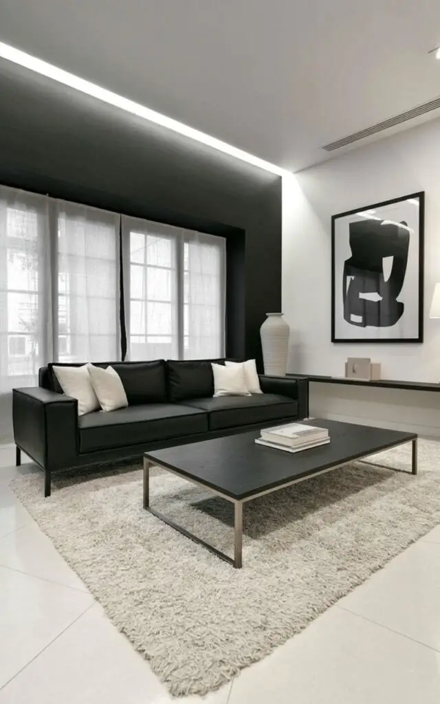 A black, low-profile sofa sits centered on a large, off-white shag rug within a living room space. The wall directly behind the sofa is painted a deep matte black, contrasting with a crisp white wall to its right. A sleek coffee table made of dark wood and a thin, brushed-metal frame rests in front of the sofa, holding a single stack of neatly arranged books. Large windows with sheer linen curtains allow soft, diffused sunlight to illuminate the room, which also features a black-framed abstract print hanging above the sofa and a tall, cylindrical white ceramic vase positioned on a nearby side table.