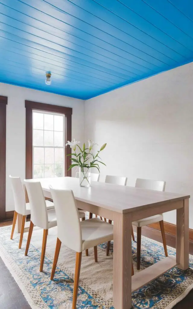 A plain, rectangular dining table with visible light wood grain sits centered in a room with a pale blue painted ceiling. Six white dining chairs with slender, tapered legs are evenly spaced around the table, each chair facing inward. Natural light streams through a window, reflecting off the smooth blue ceiling surface and creating a soft glow across the room. A simple vase containing three white lilies rests on the table's center, and a patterned blue and ivory rug lies beneath the table and chairs.