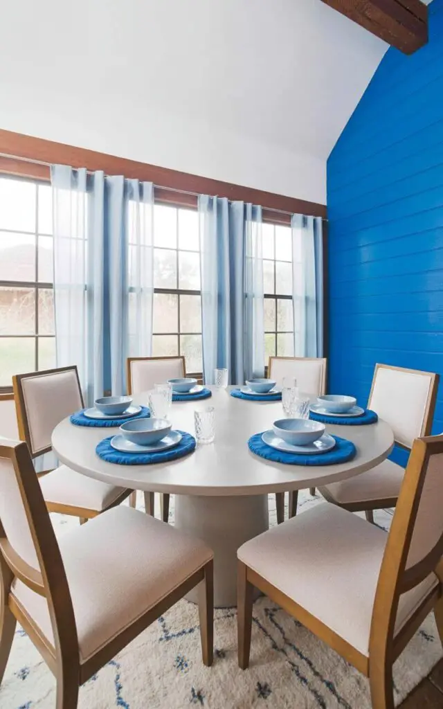 A round dining table with a smooth, light gray surface sits centered within a room, encircled by six identical dining chairs. Each chair features a straight back and armrests upholstered in a light beige linen fabric, positioned evenly around the table. The table is set with a coordinated collection of dinnerware; porcelain plates in varying shades of blue – from pale sky blue to deep navy – are layered with matching blue linen napkins and clear glass goblets. Pale blue sheer curtains hang on large windows behind the table, allowing soft natural light to illuminate the scene while a simple, white patterned rug lies beneath the table, grounding the space.