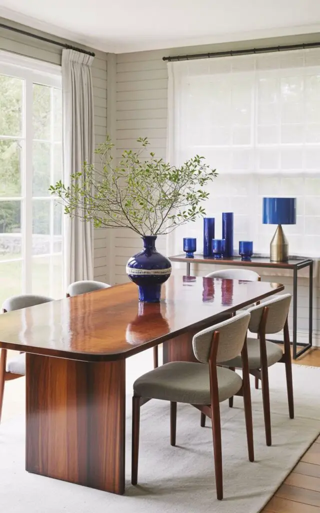 A sleek, rectangular dining table sits at the center of a blue dining room, constructed from polished dark wood with a smooth, reflective surface. A cobalt blue ceramic vase with a wide mouth and textured exterior rests on the table's surface, while six understated dining chairs upholstered in soft gray fabric surround the table with their curved backs facing inward. Cobalt blue accents appear in the form of three cylindrical pottery pieces and three matching glass tumblers placed on a narrow console table against one wall, alongside a small, brushed metal lamp with a cobalt blue lampshade. The room features light gray painted walls and a large window with white linen curtains, allowing diffused natural light to illuminate the space.