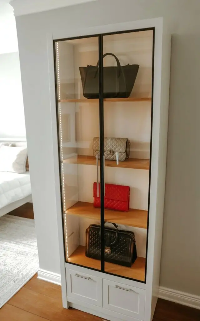 A tall, rectangular cabinet with crystal-clear glass doors stands against a pale gray bedroom wall. Inside, several handbags of varying shapes and sizes—including a structured black leather tote, a quilted beige crossbody bag, and a vibrant red clutch—rest on three adjustable oak shelves, carefully spaced to showcase each piece. Soft white LED strips are embedded along the top edge of each shelf, casting a gentle glow on the handbags and the cabinet’s brushed metal handles. The floor is covered with a light gray area rug, and a partially visible white bed frame with neutral-colored linens sits in the blurred background.