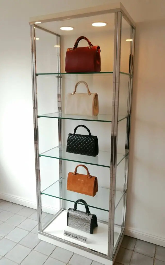 A tall glass display case with polished chrome edges stands on a light gray stone floor, presenting five handbags on individual shelves. Each shelf is softly illuminated by recessed LED lights, highlighting the textures of the handbags: one a deep burgundy leather, another a cream-colored silk, a third a black quilted fabric, a fourth a tan suede, and a fifth a metallic silver mesh. The transparent panels of the case create a sense of spaciousness, allowing unobstructed views of the bags and minimizing shadows. A small, discreet plaque near the base of the case reads "FINE LEATHERS" in a simple sans-serif font.