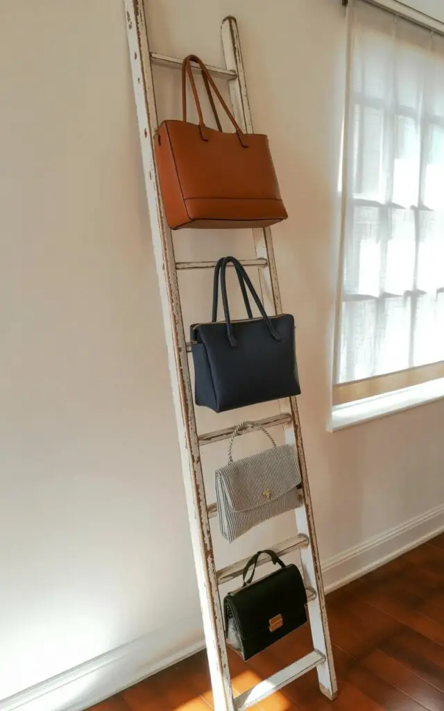 A weathered wooden ladder with evenly spaced rungs leans against a cream-colored wall with a slightly textured surface. Handbags in varying sizes and materials, including a brown leather tote, a navy blue canvas satchel, and a striped linen clutch, are draped neatly over the rungs, each bag oriented vertically. Sunlight streams through a nearby window, creating soft shadows across the wall and highlighting the natural grain of the wood. The scene takes place in a sparsely furnished room with hardwood flooring and a glimpse of a white linen curtain visible to the right of the ladder.