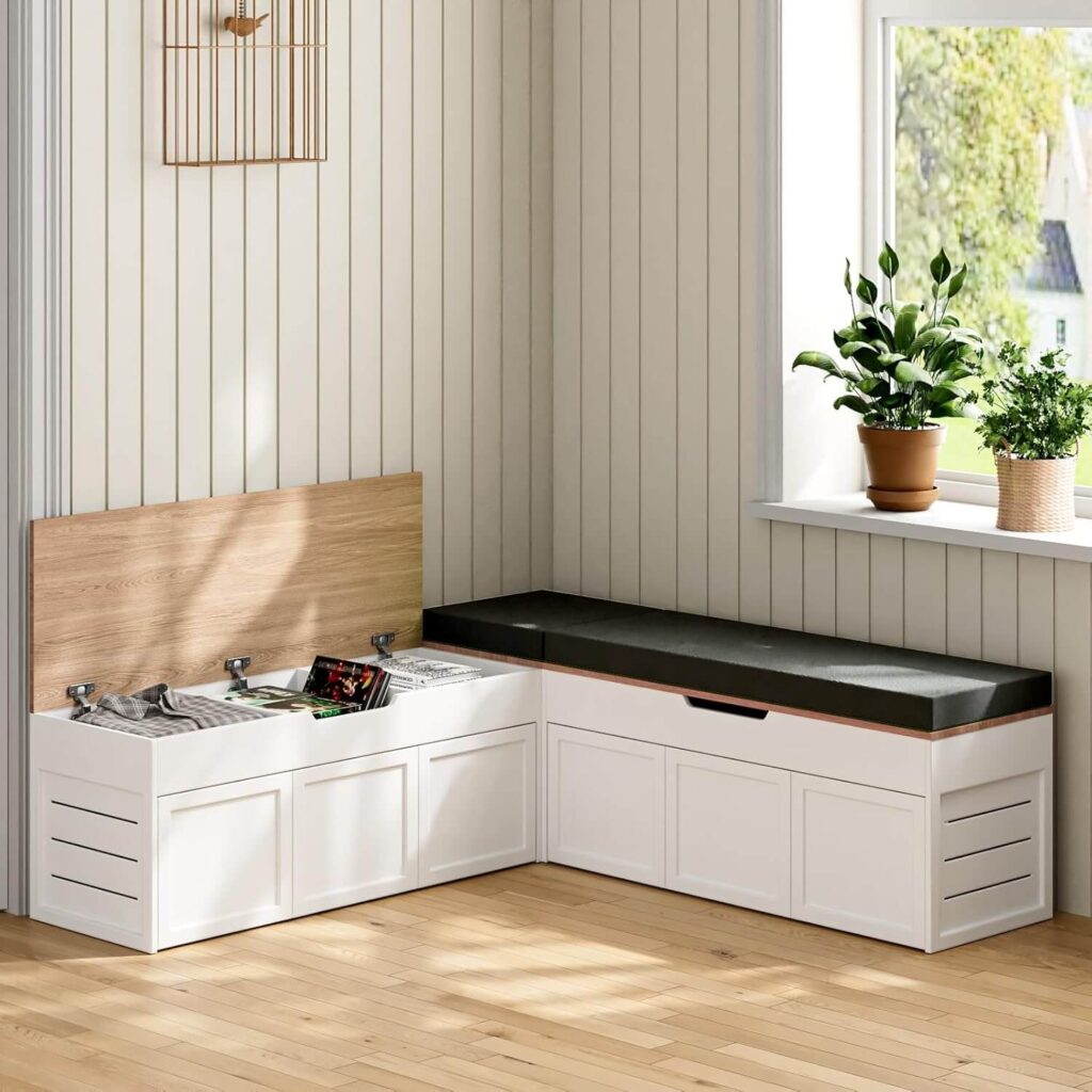 A built-in bench with a cushioned seat sits against a wall, featuring concealed storage accessed by lifting the bench cushion. Three square, light gray pillows and a folded cream-colored throw blanket are neatly arranged on the seat. A small, round wooden side table with a single open book resting on its surface is positioned to the right of the bench. The reading nook is situated within a room with pale beige walls and a large window revealing a blurred view of green trees and a clear blue sky beyond.