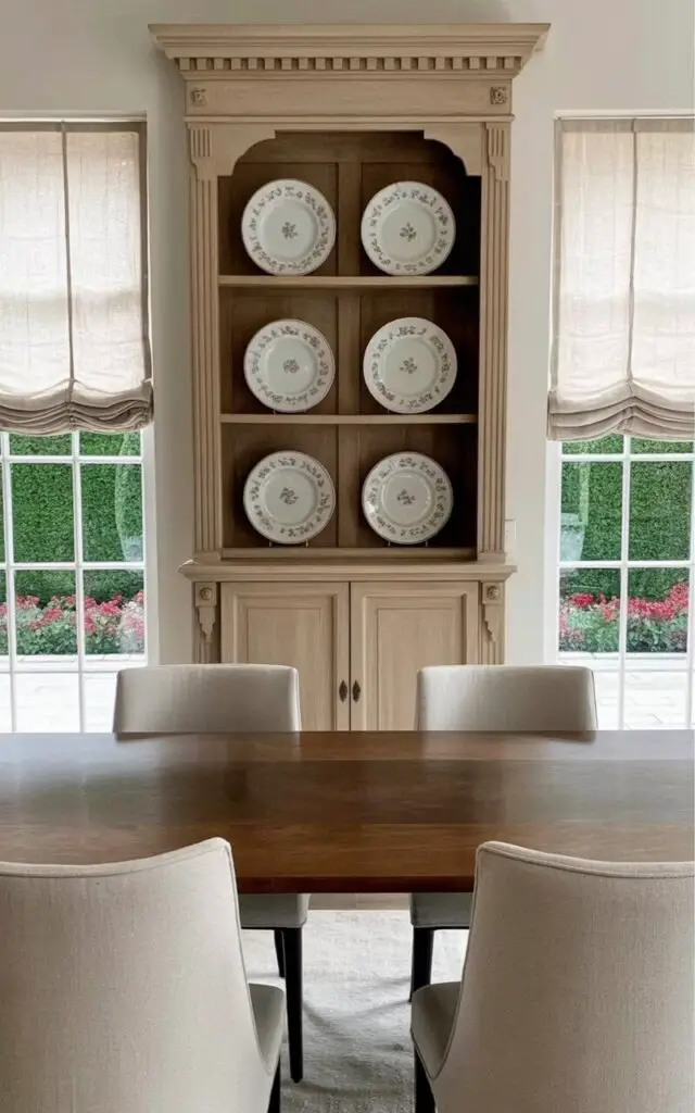 A tall wooden hutch with paneled doors and decorative crown molding stands against a pale cream-colored wall. The upper shelves of the hutch display a collection of matching porcelain dinner plates and shallow bowls with a delicate floral pattern. Beneath the hutch, a rectangular dining table made of walnut wood sits centered in the room, surrounded by four chairs upholstered in a light gray linen fabric with gently curved backs. Large windows with white linen curtains offer a view of a meticulously landscaped garden with neatly trimmed hedges and vibrant flowerbeds.