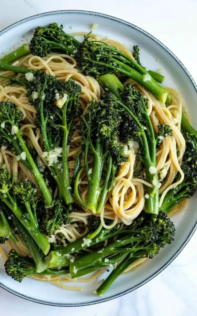 Chopped broccolini stems intertwine with long, thin strands of spaghetti pasta, scattered throughout the dish with flecks of finely minced garlic. The broccolini displays a bright green hue with a slightly tender texture, evenly distributed across the pasta. The dish is presented on a wide, circular white plate with a shallow bowl, sitting centered on a bright, clean white kitchen countertop. Soft, diffused natural light illuminates the scene, highlighting the sheen of olive oil coating the pasta and the fresh green color of the broccolini.