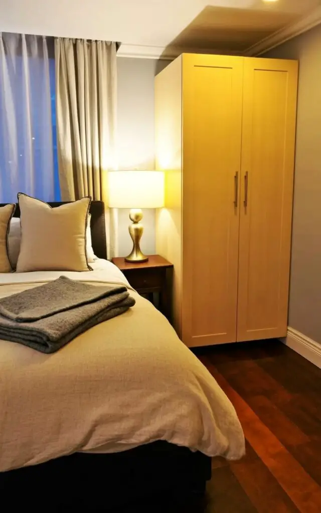 A queen-sized bed with a linen duvet cover in a light beige color and five plush pillows sits against a wall. A dark wood nightstand with a brass table lamp sits to the left of the bed, while a folded gray wool throw blanket is draped across the foot of the bed. Adjacent to the bed, a tall cabinet with a simple rectangular design and a soft, matte finish in a light oak color stands flush against the wall, featuring recessed panel detailing and brushed silver handles. The bedroom has light gray walls, wide-plank hardwood floors, and a large window with white linen curtains allows soft natural light to fill the space.