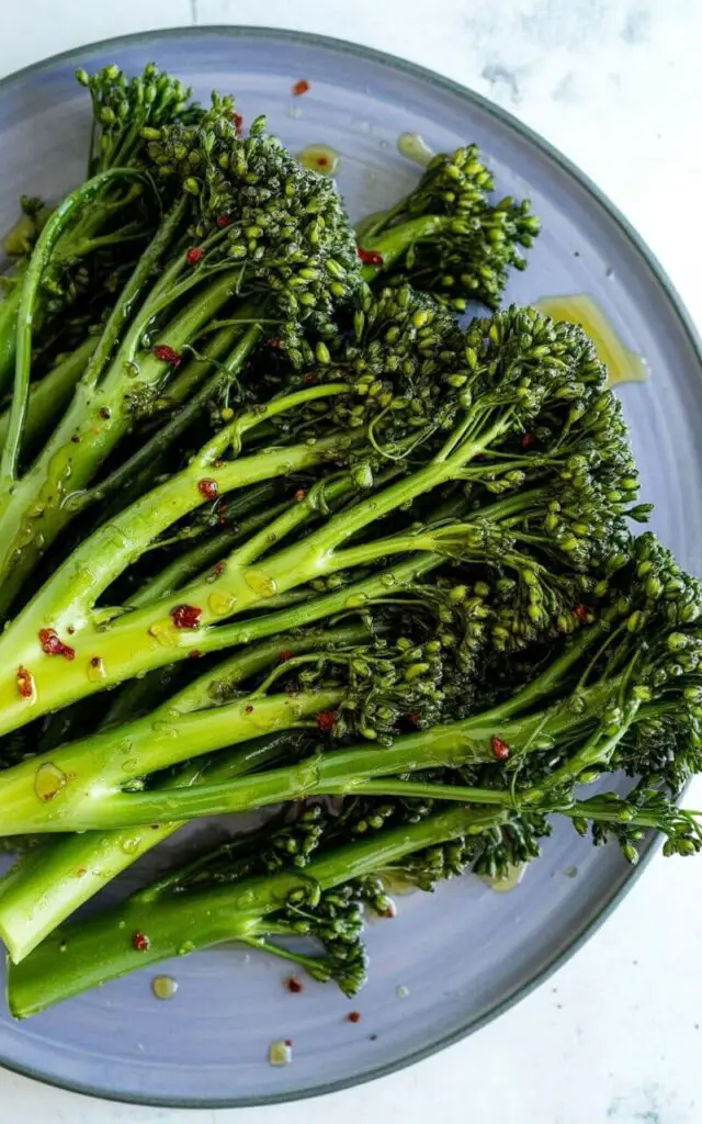 Broccolini stalks and florets are coated in glistening olive oil, with visible red chili flakes scattered across the surface. Small droplets of oil cling to the broccolini, creating highlights that accentuate its deep green color. The broccolini rests on a matte purple plate with a clean, circular rim and sits on a bright white kitchen countertop with a subtle grain texture. Soft, natural light illuminates the scene, emphasizing the contrast between the green broccolini, the red chili flakes, and the purple plate.